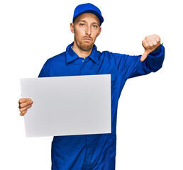 Bald man with beard wearing builder jumpsuit uniform holding empty banner with angry face, negative sign showing dislike with thumbs down, rejection concept