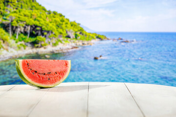 Slice of watermelon on white wooden board table and blur background of blue sea and green rocky...