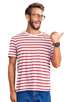 Handsome blond man with beard wearing casual clothes and glasses smiling with happy face looking and pointing to the side with thumb up.