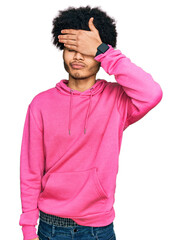 Young african american man with afro hair wearing casual pink sweatshirt covering eyes with hand, looking serious and sad. sightless, hiding and rejection concept