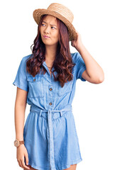 Young beautiful chinese girl wearing summer hat confuse and wondering about question. uncertain with doubt, thinking with hand on head. pensive concept.