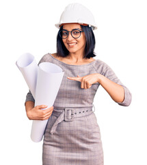 Young beautiful latin woman wearing architect hardhat and glasses holding blueprints smiling happy pointing with hand and finger