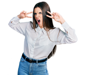 Young brunette teenager doing peace gesture close to eyes angry and mad screaming frustrated and furious, shouting with anger. rage and aggressive concept.