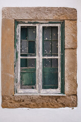 Old vintage window of an ancient stone building in Portugal. The frame is faded, glass is missing in places, and those that remain are stained with paint