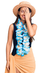 Young beautiful latin girl wearing hawaiian lei and summer hat covering one eye with hand, confident smile on face and surprise emotion.