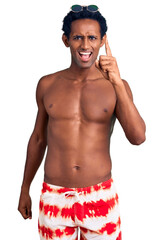 Fototapeta premium African handsome man wearing swimsuit and sunglasses pointing finger up with successful idea. exited and happy. number one.
