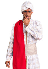 African handsome man wearing tradition sherwani saree clothes looking fascinated with disbelief, surprise and amazed expression with hands on chin