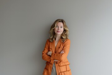 Fototapeta premium A woman with long blonde hair stands with her arms crossed in front of a gray wall, wearing a bright orange jacket.