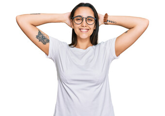 Young hispanic woman wearing casual white t shirt relaxing and stretching, arms and hands behind head and neck smiling happy