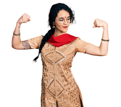 Young indian woman wearing traditional dress and glasses showing arms muscles smiling proud. fitness concept.