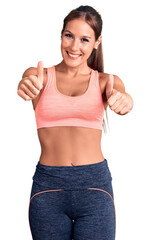 Young beautiful hispanic woman wearing sportswear approving doing positive gesture with hand, thumbs up smiling and happy for success. winner gesture.