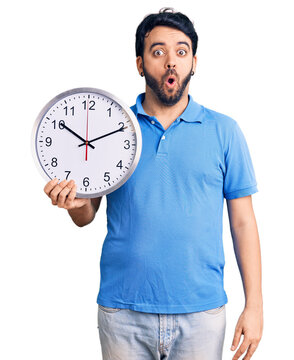 Young hispanic man holding big clock scared and amazed with open mouth for surprise, disbelief face