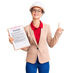 Young beautiful woman wearing architect hardhat holding clipboard with contract document surprised with an idea or question pointing finger with happy face, number one