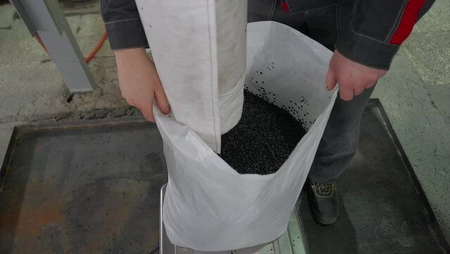 Huge Industrial Tube Pours the repurposed black plastic material pellets. Pouring lots of recycled plastic material into a white bag. Pouring manufactured plastic material into a sack.