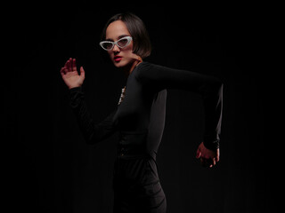 Fashionable brunette with short hair wearing white sunglasses in dark clothes on a black background  artistic light