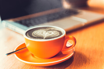 Coffee with latte art in orange cup and saucer placed on wooden table There's a laptop in the back. It represents a comfortable layout for work or study.