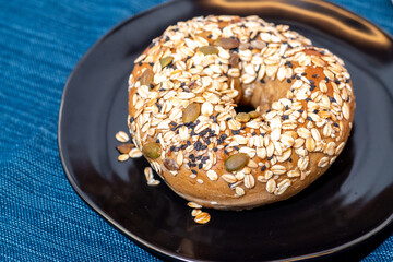 A multi-seed bagel on a black plate. The bagel is covered in a mixture of oats, sesame seeds, and pumpkin seeds.