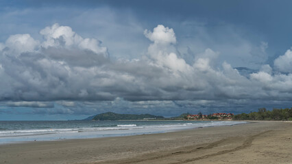 The waves of the turquoise ocean foam on the sandy beach. There are traces of a cleaning machine on the wet sand. In the distance, among the green vegetation, the hotel building. Picturesque clouds 