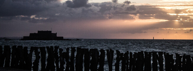 the sun is setting on the ocean behind wooden posts with clouds in the sky