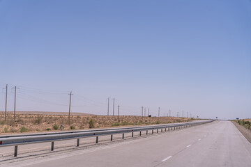 A long, empty road with a clear blue sky above. The road is lined with tall metal guard rails