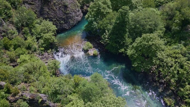 An aerial view of the Voidomatis river winding its way through the dramatic Vikos Gorge in Greece. The water is a vibrant turquoise color and the surrounding vegetation is lush and green.