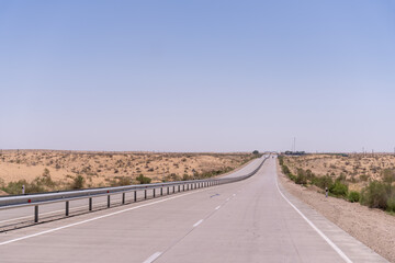 A long, empty road with a clear blue sky above. The road is surrounded by a desert landscape