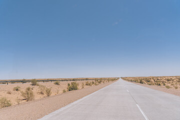 A long, empty road with a clear blue sky above. The road is surrounded by a desert landscape