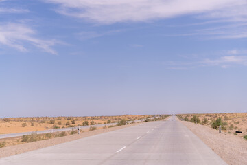 A long, empty road with a clear blue sky above. The road is surrounded by a desert landscape