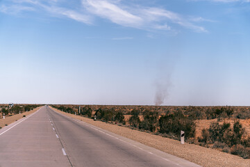 A road with a few trees in the background. The sky is clear and blue. There is a car on the road