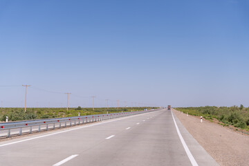 Fototapeta premium A long, empty highway with a truck driving down it. The sky is clear and blue, and there are no other vehicles on the road
