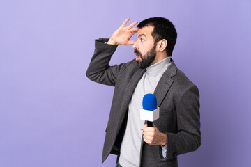 Adult reporter man with beard holding a microphone over isolated purple background with surprise...