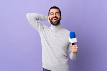 Adult reporter man with beard holding a microphone over isolated purple background laughing