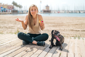 Young blonde woman with her dog holding a tartlet at outdoors making doubts gesture while lifting the shoulders