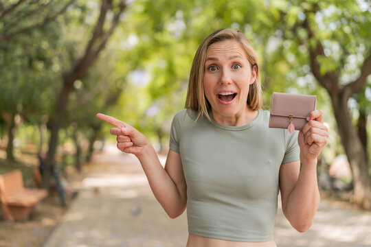 Young blonde woman holding a wallet at outdoors surprised and pointing finger to the side