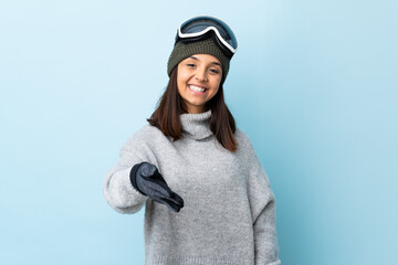 Mixed race skier girl with snowboarding glasses over isolated blue background shaking hands for closing a good deal.