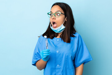 Young brunette mixed race dentist woman holding tools over isolated background doing surprise gesture while looking to the side