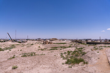 A barren desert landscape with a few buildings in the distance. The sky is clear and blue. Scene is desolate and lonely