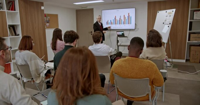 In a business training session, a professional business presenter discusses sales data on a screen to an attentive audience, showcasing expertise and engaging presentation skills.