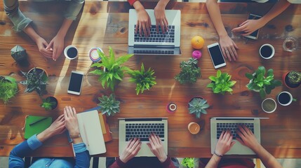 Overhead view of a team working on laptops at a wooden table with plants and coffee.