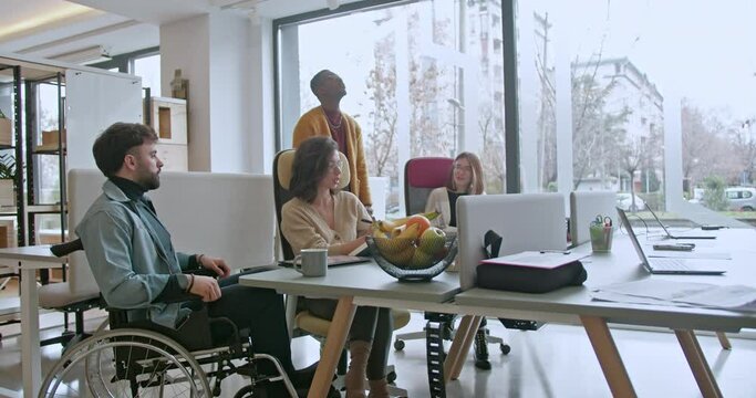 A diverse team of workers, including a person in a wheelchair, engaged in discussion at a well-lit, spacious office environment, exemplifying teamwork and inclusivity.