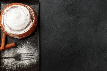 Round, delicious bun with cinnamon and powdered sugar on a black plate on a gray background
