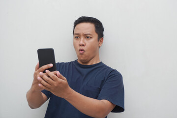 Young Asian man looking smartphone with shocked and surprised expression.  Male with blue t-shirt showing excited expression isolated on white background