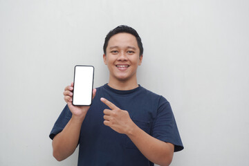 Young Asian man smiling and pointing to smartphone screen isolated on white background.  lifestyle and advertising concept. Men using blue shirt holding phone mockup