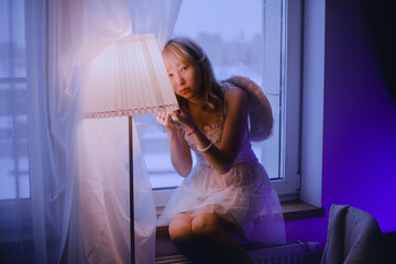 A beautiful Asian woman, dressed in white, with white wings, is sitting on a windowsill in a...