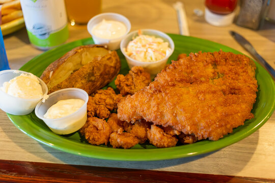 A green plate containing fried flounder, shrimp, oysters, and scallops with sides of a baked potato and coleslaw.