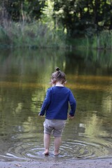 little child barefoot, seen from behind, standing in water of pond, surrounding by lush greenery. concept: summer days by water, connection with nature, innocence and discovery, childhood adventures.