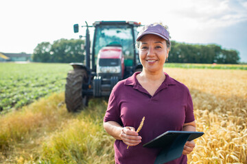 Portrait of a smiling female farmer in front of tractor with tablet in wheat field