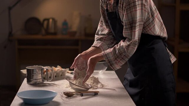 Female baker pouring flour into a metal sifter with hands, carefully preparing baking ingredients on a rustic kitchen table.