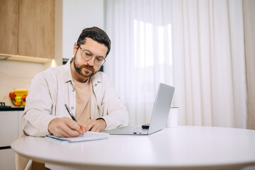Busy caucasian confident man freelancer writing information on notepad in home office working with laptop