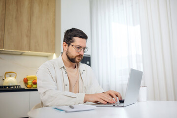 caucasian male typing on personal laptop and working remotely in home space. Positive bearded man freelancer dressed in casual clothes and glasses on kitchen.